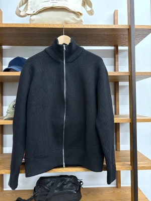 Svart stickad tröja Maison Margiela - Svart tröja från Maison Margiela med ribbad stickning och dragkedja framtill. Tröjan har högt krage och de ikoniska vita sömmarna i nacken. Perfekt för dig som gillar minimalistisk och clean stil.