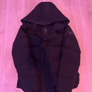 Svart pufferjacka från Canada Goose - Svart pufferjacka från Canada Goose med huva och klassisk logga på ärmen. Jackan har quiltad design, dragkedja och knappar framtill. Perfekt för kalla dagar med sin tjocka och isolerande fyllning. Snygg och stilren look som funkar till alla dina vinteroutfits.