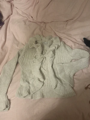 Beige stickad kofta med volang från Bershka - Superfin beige stickad kofta från Bershka med volangkant och knyt framtill. Koftan har långa ärmar och ribbad struktur, perfekt för lager-på-lager och en mysig vibe.