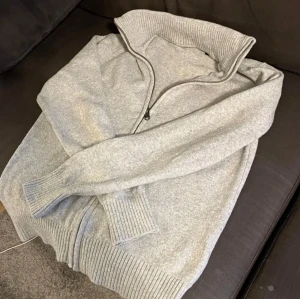 Ljusgrå stickad kofta med dragkedja - Ljusgrå stickad kofta med hög krage och hel dragkedja framtill. Tröjan har ribbade muddar vid ärmslut och nederkant, vilket ger en schysst passform. Perfekt för lager-på-lager och chill vardagsstil.