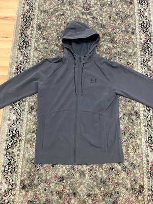 Grå vindjacka från Under Armour M - Säljer en grå vindjacka från Under Armour med huva och hel dragkedja. Jackan har två sidofickor, justerbar dragsko i huvan och meshfoder på insidan. Perfekt för blåsiga dagar och sportiga outfits.