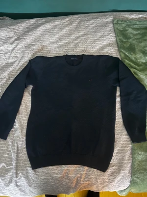 Svart stickad tröja Tommy Hilfiger - Svart stickad tröja från Tommy Hilfiger med diskret logga på bröstet. Tröjan har rund hals, ribbade muddar och långärmad passform. Materialet är mjukt och strukturerat, perfekt för dig som gillar klassisk stil med en modern touch. Liten i stl passar mig som är 163 men börjar bli liten