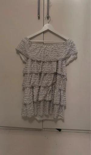 Vit prickig volangklänning - Supersöt kort volangklänning i vitt med svarta prickar. Klänningen har flera lager volanger och en offshoulder-ringning som ger en romantisk vibe. Tillverkad i ett lätt, luftigt material som känns perfekt för varma dagar. Prickarna ger en lekfull touch och volangerna gör den extra speciell.