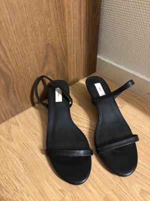 Svarta klackar från Nelly - Stilrena svarta klackar från Nelly med smala remmar och låg klack. Skorna har en rund tå och är tillverkade i syntetmaterial. Perfekt för dig som gillar minimalistisk design och vill ha en enkel men snygg look. Använda någon enstaka gång!🖤