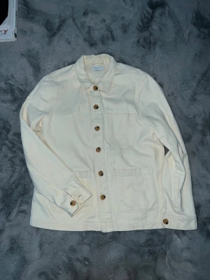 Vit/krämig overshirt från Studio Total - Funkar även för S. vit/cream overshirt från Studio Total i bomull. Jackan har klassisk krage, bruna knappar framtill och tre praktiska fickor. Perfekt lager-på-lager-plagg med enkel och clean look.