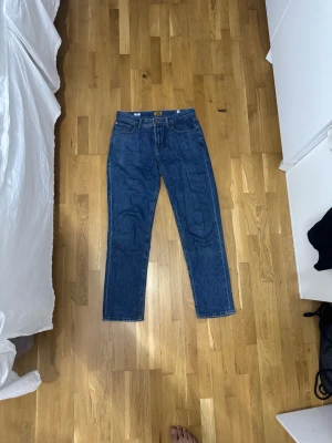 Blå jeans från Jack & Jones - Säljer ett par klassiska blå jeans från Jack & Jones med rak passform och fem fickor. Jeansen har normal midja, snygga detaljer med knapp och dragkedja framtill samt robust denimkänsla. Perfekt för dig som gillar enkel och tidlös stil.