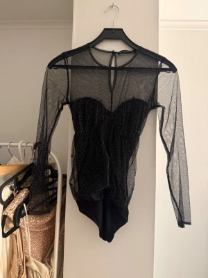 Svart mesh body med glitterprickar - Svart mesh body med långa ärmar och glittriga små prickar över hela tyget. Toppen har en snygg sweetheart-urringning och knäppning i nacken. 