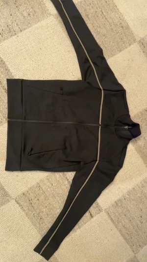 Svart vindjacka från BOSS med beige detaljer - Svart tröja från BOSS med beige ränder längs ärmar och bröst. Tröjan har hög krage, hel dragkedja och två fickor med dragkedja. Materialet är stretchigt och känns sportigt, perfekt för en chill och stilren look.  är köpt på johnells för 2199 aldrig använt för är inte min still så säljer den för 1100 för vill bli av med den kan gå ner lite mer om de är ett rimligt pris 