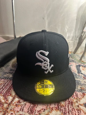 Chicago White Sox svart keps New Era - Svart 59FIFTY keps från New Era med Chicago White Sox-logga broderad i vitt framtill. Snygg platt skärm med grå undersida och broderad text på sidan: 'Chicago, Illinois established 1900 Windy City!'. Perfekt för dig som gillar baseball och streetwear.