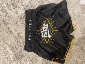 Svarta Fairtex träningsshorts - Snygga svarta träningsshorts från Fairtex med gul kant och stort tryck på framsidan. Shortsen har bred resår i midjan och är perfekta för kampsport eller gym. Materialet är glansigt och lätt, vilket ger bra rörelsefrihet. Det år storlek Xl men sitter lite mer som L 