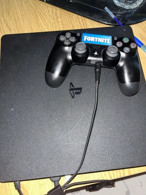 PlayStation 4 med DualShock 4 Fortnite Edition - Snygg PlayStation 4 med original DualShock 4-kontroll, dekorerad med Fortnite-märke. Konsolen och kontrollen är i bra begagnat skick med mindre ytliga repor. Perfekt för dig som vill spela populära spel som Fortnite direkt! Alla kablar medföljer.