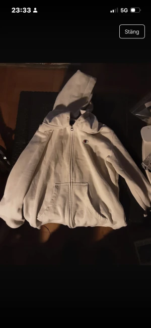 Vit hoodie från Champion med dragkedja - Säljer en vit hoodie från Champion med huva och praktisk dragkedja framtill. Tröjan har två stora fickor och den klassiska Champion-loggan på bröstet. Perfekt för chill dagar och streetstyle. Passar dig som gillar enkel och clean look.