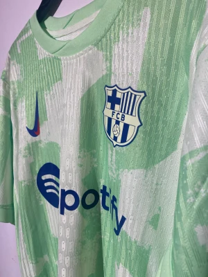 FC Barcelona Lamine Yamal bortatröja - Säljer en FC Barcelona bortatröja från Nike med Lamine Yamal 19 på ryggen. Tröjan är ljusgrön med vitt mönster, har korta ärmar och är tillverkad i ett lätt, ventilerande funktionsmaterial. Tryck med Spotify-logga och klubbmärke på bröstet. Nu under black friday veckan för endast 349 kr🔥🔥