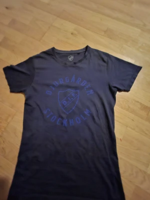 Djurgården svart t-shirt S - Svart t-shirt i storlek S med Djurgården IF Stockholm-tryck i blått på bröstet och DIF-logga i nacken. Klassisk rund hals och korta ärmar. Perfekt för dig som vill visa ditt stöd för Djurgården. Materialet är mjuk bomull.