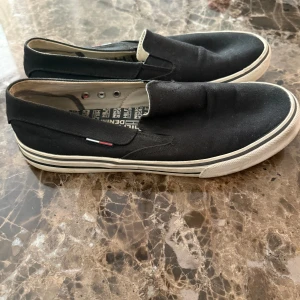 Svarta slip-on sneakers Tommy Hilfiger - Svarta slip-on sneakers från Tommy Hilfiger med vit sula och diskret logga på sidan. Skorna är i canvasmaterial och har en klassisk, låg profil med rund tå och platt sula. Perfekta för en avslappnad och stilren look.