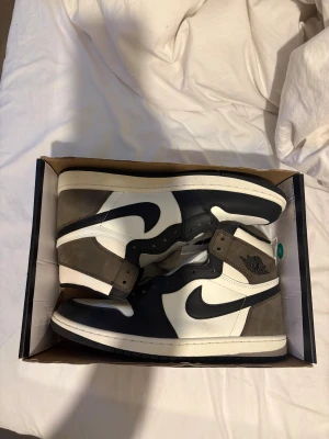 Nike Air Jordan 1 High Mocha - STORLEK 46! Säljer ett par Nike Air Jordan 1 High med svart, vit och brun färgkombination. Skorna har klassisk hög siluett, svart swoosh och detaljer i mocka och skinn. Ikonisk Jordan Wings-logga på sidan och perforerad tå. Extra vita skosnören medföljer. Misstänker att de är ej äkta då jag köpte på plick.