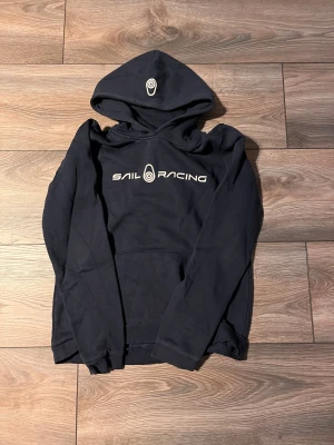 Mörkblå Sail Racing hoodie med tryck - Snygg mörkblå hoodie från Sail Racing med vit logotyp på bröstet och på huvan. Tröjan har en klassisk känguruficka framtill och långa ärmar. Mjuk insida och ribbade muddar vid ärmslut och nederkant. Perfekt för chill eller häng med kompisar. Storlek 176