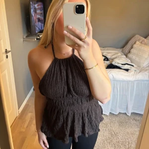 Brun halterneck topp med öppen rygg från Gina tricot - Supersnygg brun halterneck topp från Gina Tricot med omlott och knyt i ryggen. Toppen har en crinklad struktur, volangkant nedtill och är ärmlös med öppen rygg. Perfekt att styla med jeans eller kjol för en trendig look.