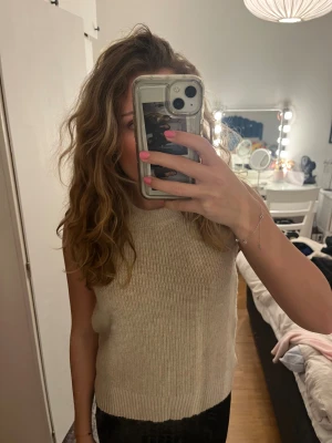 Beige stickad ärmlös topp - Säljer en beige stickad topp med rund hals och ribbad struktur. Modellen är ärmlös och har en mjuk, lite loose passform som är enkel att styla. Perfekt för dig som gillar neutrala färger och enkel design.