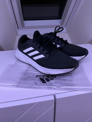 Svarta adidas träningsskor  - Svarta Adidas träningsskor med klassiska vita ränder på sidorna och vit, tjock sula. Ovandelen är i mesh-liknande syntetmaterial och skorna har snörning. Perfekta för träning eller en sportig look. Adidaslogga på sulan och sidan.