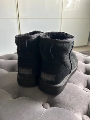 Svarta UGG boots i mocka - Mysiga svarta UGG boots med rund tå och platt sula. Utsidan är i mjuk mocka och insidan är fodrad med fluffig päls för extra värme. Perfekta för kalla dagar och har en klassisk, enkel design med UGG-logga bak på hälen.