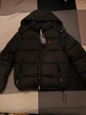 Svart dunjacka Polo Ralph Lauren - Svart dunjacka från Polo Ralph Lauren med huva och klassisk röd logga på bröstet. Jackan har dragkedja framtill och två dragkedjeförsedda fickor. Puffig modell som håller dig varm och snygg under kalla dagar. Perfekt för dig som gillar stilren och sportig look.