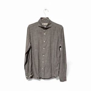 Grå skjorta från Morris i bomull - Stilren grå skjorta från Morris med klassisk krage och vita knappar. Tillverkad i italienskt bomullstyg från Albini, vilket ger en mjuk och lyxig känsla. Skjortan har lång ärm och en diskret, melerad struktur som ger en modern look.