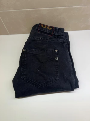 Svarta jeans från Dondup - Svarta jeans från Dondup med klassisk femficksdesign och silverfärgade nitar. Snyggt D-emblem på bakfickan och diskret röd detalj på framsidan. Jeansen har rak passform och är tillverkade i slitstarkt bomullsmaterial. Perfekt för dig som gillar stilrena och tidlösa jeans.