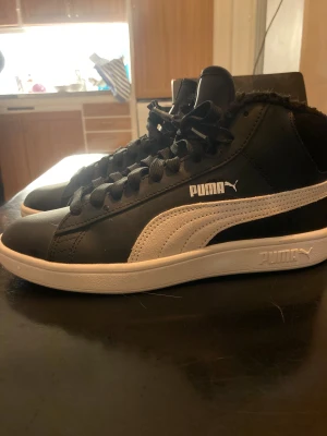 Svarta höga sneakers från Puma - Säljer ett par svarta höga sneakers från Puma med vit sula och vit klassisk Puma-logga på sidan. Skorna har snörning, rund tå och är tillverkade i syntetmaterial med mjukt foder runt ankeln. Perfekta för dig som gillar streetstyle och vill ha en clean look. Den passar in med allt och håller värmen länge!