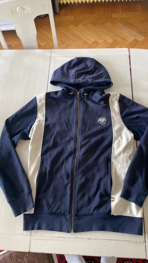 Blå och vit hoodie Roland Garros L - Snygg hoodie från Roland Garros i storlek L.  Nypris är runt 900kr, kom inte riktigt till användning, så är i  bra skick. För mer frågor eller bilder är det bara att skriva!