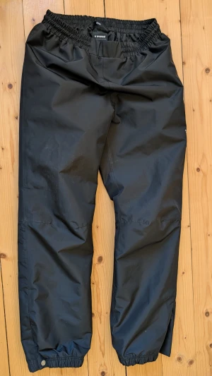 Svarta regnbyxor från Everest - Svarta regnbyxor från Everest med elastisk midja och justerbara benslut med knapp. Perfekta för regniga dagar och utomhusaktiviteter. Materialet är slitstarkt och vattenavvisande syntet, vilket gör dem grymt praktiska när det är blött och kallt ute.