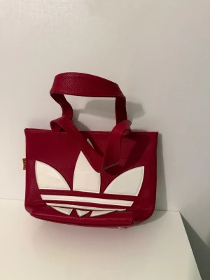 Röd axelväska från Adidas - Snygg röd axelväska från Adidas med stort vitt logotryck på framsidan. Väskan har två rejäla handtag och en praktisk dragkedja på baksidan. Materialet är skinnimitation med struktur och väskan har en modern, boxig form.