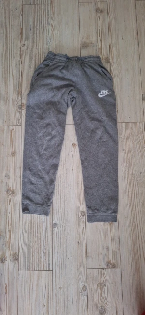Gråa mjukisbyxor från Nike - Gråa mjukisbyxor från Nike med vit logga på vänster lår. Byxorna har resår i midjan och vid bensluten, samt sidofickor. Perfekta för chill eller träning, tillverkade i mjukt bomullsmaterial.