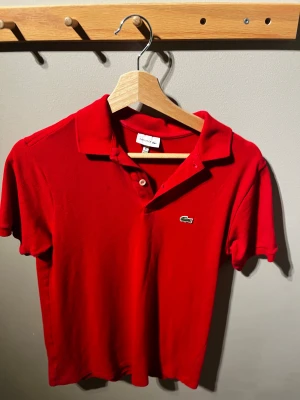 Röd pikétröja från Lacoste - Säljer en klassisk röd pikétröja från Lacoste med korta ärmar och krage. Tröjan har två knappar vid halsen och den ikoniska gröna krokodilloggan broderad på bröstet. Materialet är mjuk bomull som andas och passar perfekt till en chill stil.