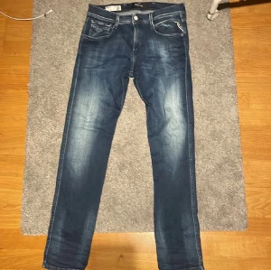 Replay Anbass jeans blå, storlek 30/32 - Snygga Replay Anbass jeans i mörkblå tvätt med slitningar och kontrastsömmar. Modellen har fem fickor, dragkedja och knapp framtill. Jeansen är raka och har en modern passform som sitter riktigt bra. Perfekt för dig som gillar stilrena och trendiga jeans. Jeansen är i storlek 30 i midjan och 32 i längden