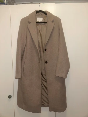 Beige kappa med knappar från HM - Stilren beige kappa med klassisk krage och två mörka knappar framtill. Lång modell med raka ärmar och diskreta fickor i sidorna. Perfekt för dig som gillar en clean och tidlös look.