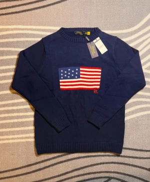 Mörkblå stickad tröja Polo Ralph Lauren - Snygg mörkblå stickad tröja från Polo Ralph Lauren med amerikansk flagga broderad på bröstet och RL-detalj i rött. Tröjan har rund hals och ribbade muddar vid ärmslut och nederkant. Perfekt för dig som gillar klassisk stil med en twist.