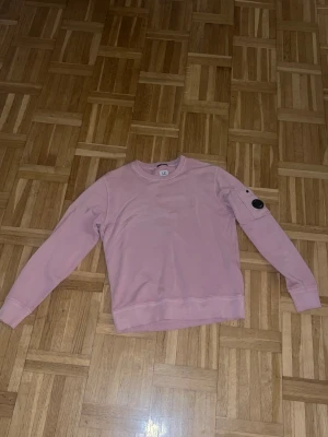 Rosa sweatshirt från C.P. Company - Snygg rosa sweatshirt från C.P. Company med klassisk rund hals och lång ärm. Tröjan har en ikonisk svart linsdetalj på ärmen och ribbade muddar vid ärmslut och nederkant. Tillverkad i mjuk bomull som är resist dyed för extra cool look.
