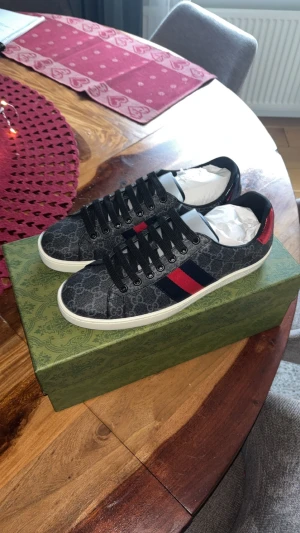 Gucci ace sneakers  - Snygga Gucci sneakers i mörkgrått med klassiskt GG-mönster och vita sulor. Skorna har svarta snören, röd och marinblå rand på sidan samt metallic-detaljer i rött och svart på hälen med Gucci-logga. Materialet är canvas och skinn, med rund tå och platt sula.