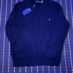 Svart stickad tröja Polo Ralph Lauren - Snygg svart stickad tröja från Polo Ralph Lauren med klassiskt kabelstickat mönster och röd broderad logga på bröstet. Rund hals och långa ärmar med ribbade muddar. Perfekt för dig som gillar stilrena och tidlösa plagg. Priset kan prutas