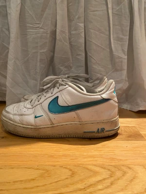Nike Air Force 1 vit med turkos swoosh - Nike Air Force 1 sneakers i vitt skinn med turkos swoosh och detaljer. Klassisk låg modell med snörning och platt sula. Snygg retrostil med tydlig Air-logga på hälen och turkosa accenter som sticker ut.