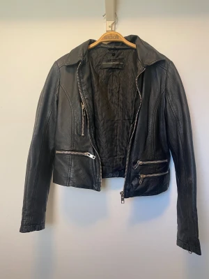 Svart skinnjacka med dragkedjor från Bik Bok - Snygg svart skinnjacka från Bik Bok med flera silvriga dragkedjor. Jackan har klassisk krage, figurnära passform och långa ärmar. Väl använd men i väldigt fint skick! 