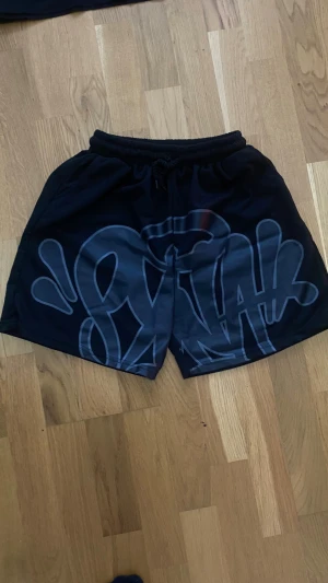 Svarta shorts från Syna World - Svarta shorts i storlek S med ett stort grafiskt tryck framtill i grått. De har elastisk midja med snörning för skön passform och är gjorda i polyester. Perfekta för chill dagar eller streetwear-stil. Priset är lågt av en anledning.