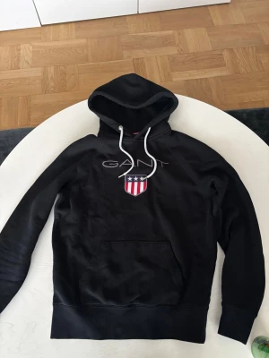 Svart hoodie från GANT med logga - Svart hoodie från GANT med vit snörning i huvan och stor broderad logga på bröstet. Tröjan har en klassisk känguruficka framtill och mjukt bomullsmaterial. Snyggt emblem med stjärnor och ränder ger en sportig vibe. Perfekt för chill dagar.