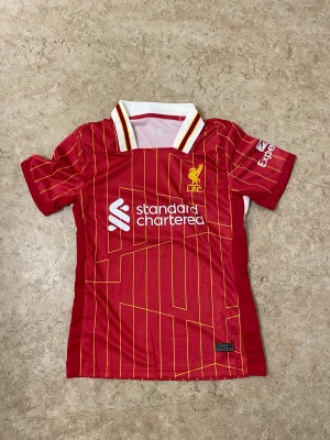 Liverpool FC röd matchtröja - Snygg röd Liverpool FC fotbollströja med gula och vita detaljer, klassisk krage och tunna gula ränder. Tryck med Standard Chartered på bröstet och klubbmärke i gult. Tillverkad i lätt polyester som andas, perfekt för träning eller match.