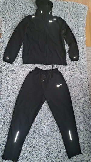 Svart Nike träningsset med reflex - Nike träningsset i svart med jacka och byxor. Jackan har huva, dragkedja och reflexdetaljer på axlar och ärmar. Byxorna har snörning i midjan, reflexdetaljer vid benslut och Nike-logga på låret. Materialet är lätt och syntetiskt, perfekt för aktiva dagar. Helt ny me etikettet på