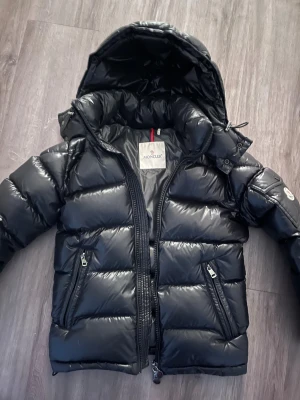 Svart Moncler Maya stl xs - Svart Moncler med glansig finish, huva och två snedställda fickor med dragkedja. Jackan har klassisk Moncler-logga på ärmen och quiltad design. Perfekt för kalla dagar. Mycket fint skick. NFC och scan fungerar. 