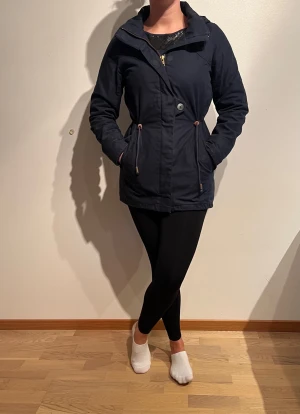 Marinblå jacka, Elvine.  - Material: Yttermaterial: 65% polyester, 35% bomull. Foder: 100% polyester. Fyllning: 100% polyester   Tvättråd: 40° Övrigt: Avtagbar luva. Magnet längst med dragkedja och fickor