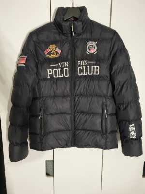 Svart pufferjacka Vinson Polo Club - Svart pufferjacka från Vinson Polo Club med broderade loggor och flaggor på bröstet och ärmen. Jackan har dragkedja framtill, två sidofickor med dragkedja och hög krage. Materialet är syntet och jackan är quiltad för extra värme.🩷den är i storlek S