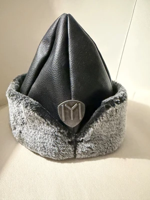 Svart pälsfodrad hatt med emblem( osmaklig) - Unik svart hatt med spetsig form, tillverkad i skinnimitation och fodrad med mjuk grå päls runt kanten. Framtill sitter ett metallfärgat emblem med YM-logga. Perfekt för dig som vill sticka ut med en cool accessoar.